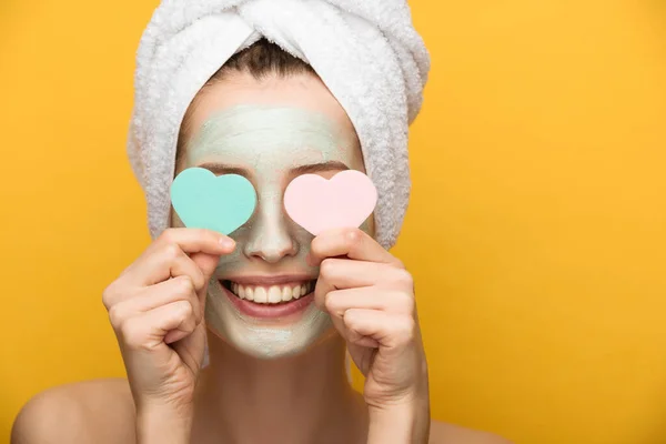 Sorridente ragazza con maschera viso nutriente che copre gli occhi con spugne cosmetiche a forma di cuore su sfondo giallo — Foto stock