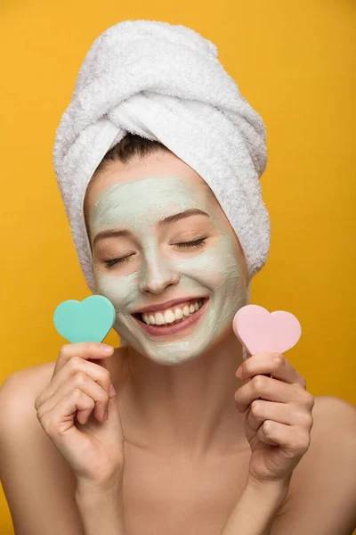 Chica alegre con mascarilla facial nutritiva sosteniendo esponjas cosméticas en forma de corazón con los ojos cerrados aislados en amarillo - foto de stock