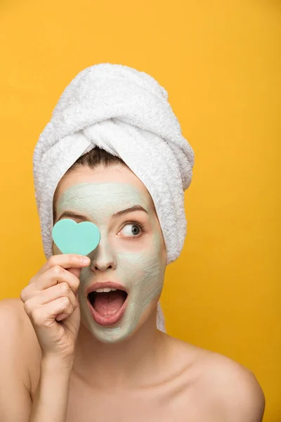 Surprised girl with nourishing facial mask covering eye with heart-shaped cosmetic sponge isolated on yellow — Stock Photo