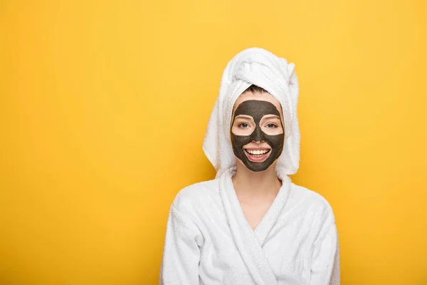 Smiling girl with facial clay mask and towel on head looking at camera on yellow background — Stock Photo