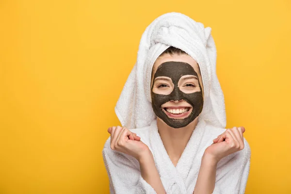 Chica feliz con máscara de arcilla facial y toalla en la cabeza sosteniendo puños apretados y mirando a la cámara aislada en amarillo - foto de stock