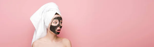 Plan panoramique de fille choquée avec masque d'argile faciale et serviette sur la tête regardant loin isolé sur rose — Photo de stock