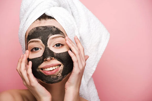 Sorridente ragazza con maschera di argilla facciale toccare il viso e guardando la fotocamera isolata su rosa — Foto stock