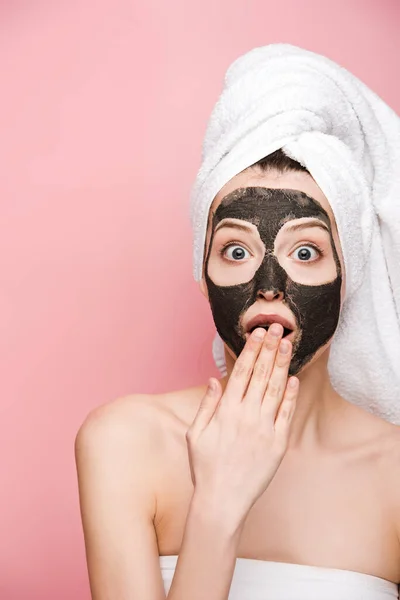 Shocked girl with clay mask on face covering mouth with hand and looking at camera isolated on pink — Stock Photo