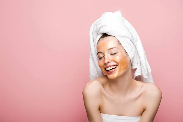 Cheerful girl with facial mask and towel on head smiling with closed eyes isolated on pink — Stock Photo