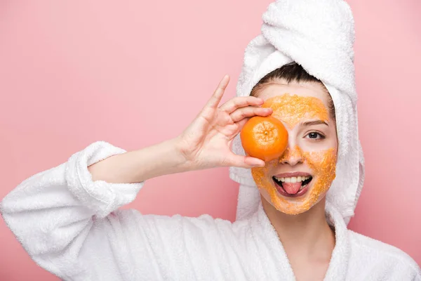 Menina alegre com máscara facial cítrica cobrindo o olho com tangerina e salientando a língua isolada no rosa — Fotografia de Stock