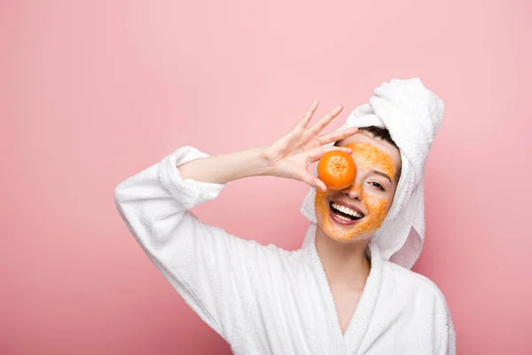 Веселая девушка с цитрусовыми масками для лица, закрывающая глаза мандарином на розовом фоне — стоковое фото