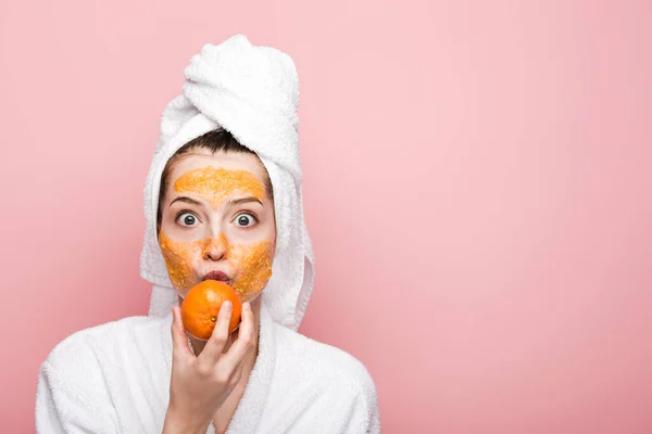 Excited girl with citrus facial mask holding tangerine near mouth isolated on pink — Stock Photo