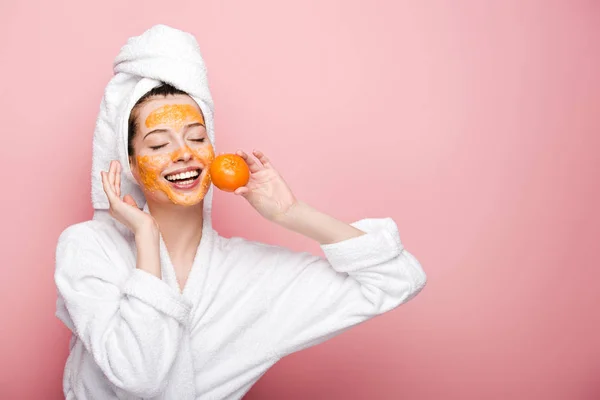 Glückliches Mädchen mit Zitrusgesichtsmaske, die Mandarine in Gesichtsnähe auf rosa Hintergrund hält — Stockfoto