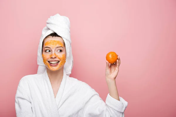 Happy girl with citrus facial mask looking at tangerine isolated on pink — Stock Photo