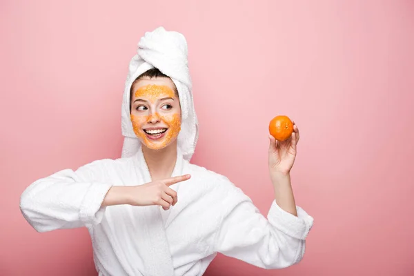 Menina alegre com máscara facial cítrica apontando com o dedo para tangerina no fundo rosa — Fotografia de Stock