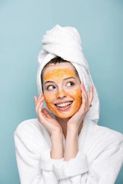 Menina sorridente com máscara facial cítrica tocando rosto enquanto olhando para longe isolado em azul — Fotografia de Stock