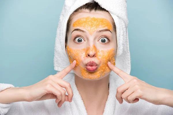 Aufgeregtes Mädchen mit zitrusfarbener Gesichtsmaske, das Gesicht berührt, während es isoliert auf blau in die Kamera schaut — Stockfoto