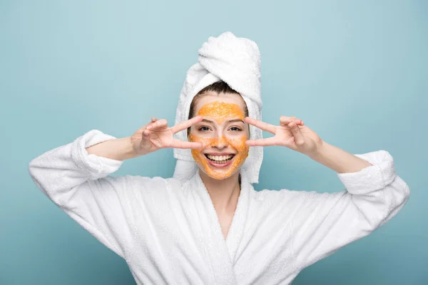 Gaie fille avec des agrumes masque facial montrant des gestes de victoire sur fond bleu — Photo de stock