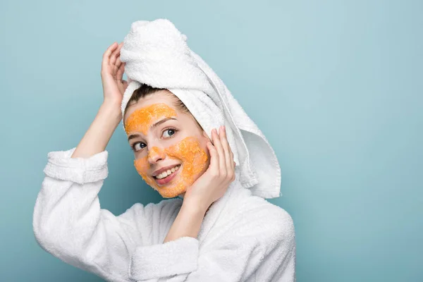 Lächelndes Mädchen mit zitrusfarbener Gesichtsmaske, die Gesicht und Handtuch auf dem Kopf berührt, während sie auf blauem Hintergrund wegschaut — Stockfoto
