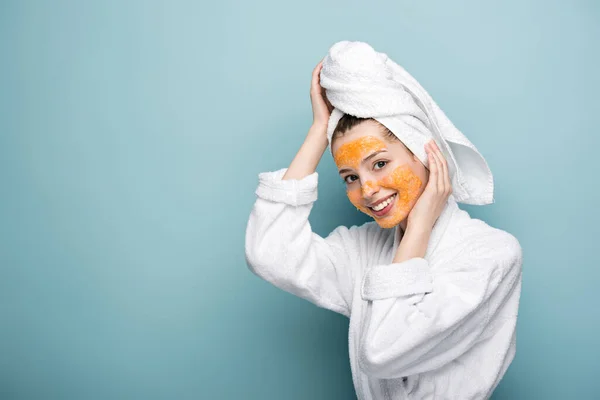 Fille souriante avec des agrumes masque facial serviette touchante sur la tête sur fond bleu — Photo de stock