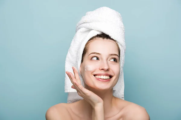 Feliz chica atractiva aplicando mascarilla facial hidratante mientras mira hacia otro lado aislado en azul - foto de stock