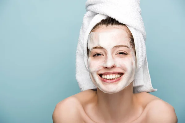 Cheerful girl with moisturizing facial mask smiling at camera isolated on blue — Stock Photo