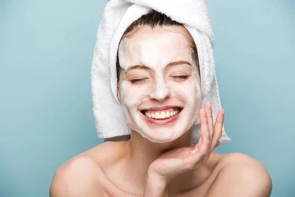 Menina feliz com máscara facial hidratante tocando o rosto com os olhos fechados isolados no azul — Fotografia de Stock