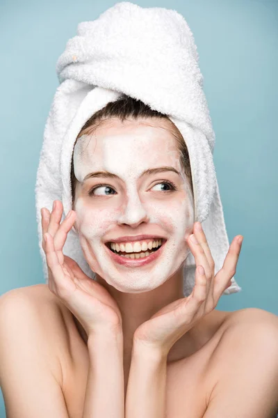 Chica feliz con mascarilla facial hidratante tocando la cara y mirando hacia otro lado aislado en azul - foto de stock