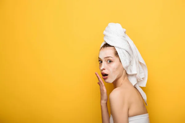 Shocked girl with moisturizing facial mask holding hand near mouth and looking at camera isolated on yellow isolated on yellow — Stock Photo