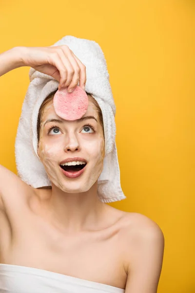 Excitada chica con mascarilla facial hidratante mirando esponja cosmética aislado en amarillo - foto de stock