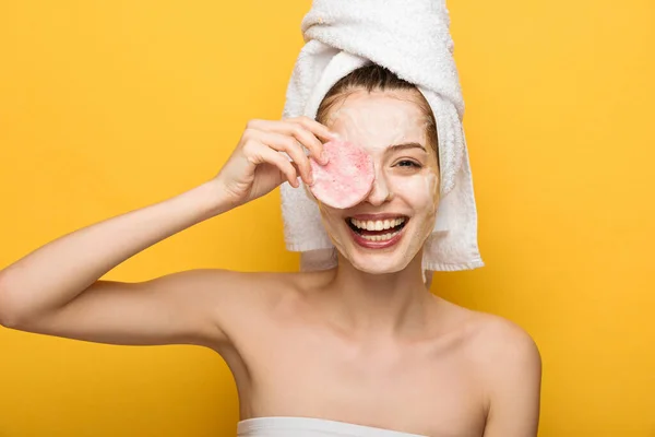 Cheerful girl with moisturizing facial mask covering eye with cosmetic sponge on yellow background — Stock Photo