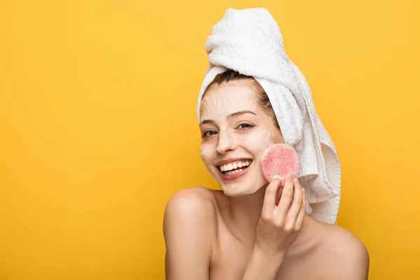 Menina feliz com máscara facial hidratante olhando para esponja cosmética isolada no amarelo — Fotografia de Stock