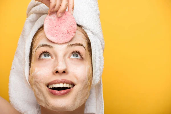 Chica alegre con mascarilla facial hidratante mirando esponja cosmética aislado en amarillo - foto de stock