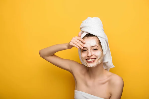 Menina alegre com máscara facial hidratante tocando o rosto e olhando para a câmera no fundo amarelo — Fotografia de Stock