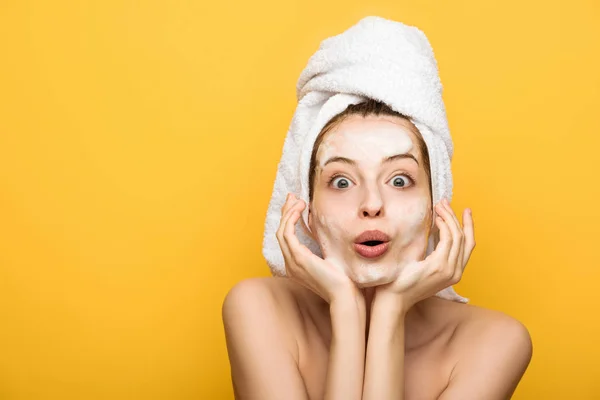 Chica sorprendida con mascarilla facial hidratante cogida de la mano cerca de la cara mientras mira la cámara aislada en amarillo - foto de stock