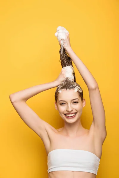 Fille heureuse regardant la caméra tout en lavant les cheveux longs sur fond jaune — Photo de stock