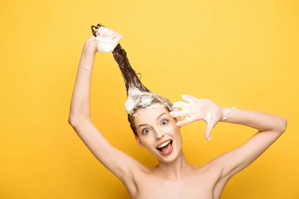 Glückliches Mädchen zeigt seifige Hand beim Waschen langer Haare auf gelbem Hintergrund — Stockfoto