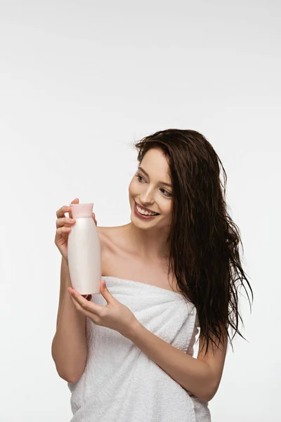 Chica sonriente mirando hacia otro lado mientras sostiene la botella de champú aislado en blanco - foto de stock
