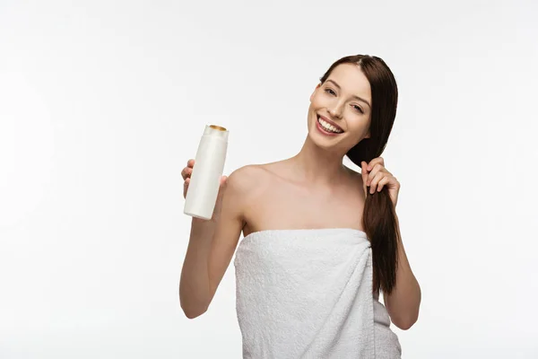 Cheerful girl touching long shiny hair while holding shampoo isolated on white — Stock Photo