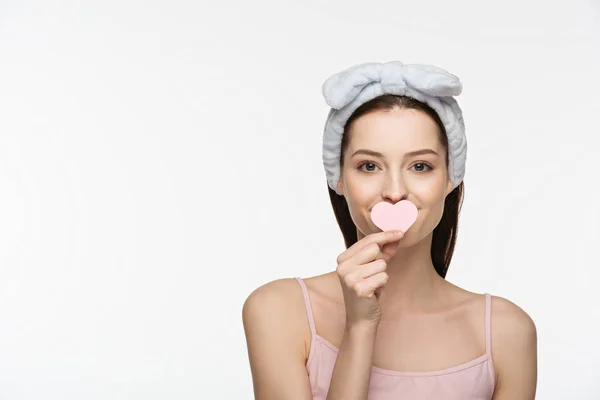 Fröhliches Mädchen, das den Mund mit einem herzförmigen Kosmetikschwamm bedeckt, während es isoliert auf weiß in die Kamera blickt — Stockfoto