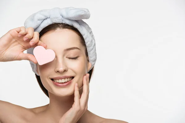 Happy girl covering eye with heart-shaped cosmetic sponge while touching face isolated on white — Stock Photo