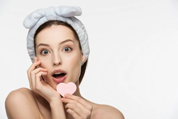 Shocked girl holding heart-shaped cosmetic sponge while looking at camera isolated on white — Stock Photo