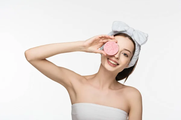 Happy girl covering eye with cosmetic sponge while looking at camera isolated on white — Stock Photo