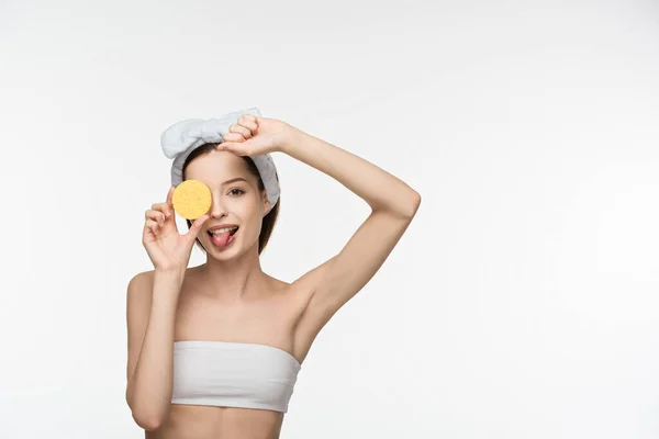 Fröhliches Mädchen, das das Auge mit Kosmetikschwamm bedeckt und die Zunge isoliert auf weißem Grund ausstreckt — Stockfoto