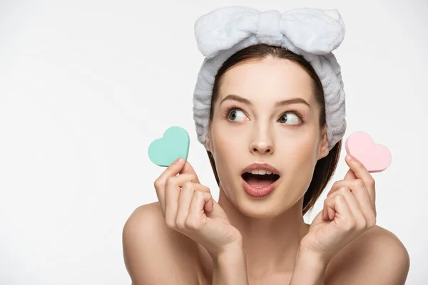Shocked girl holding heart-shaped cosmetic sponges while looking away isolated on white — Stock Photo