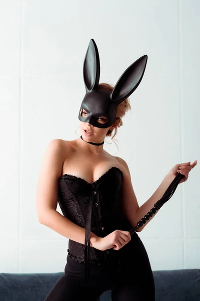 Dominant young woman in bunny mask and corset holding flogging whip and looking away — Stock Photo