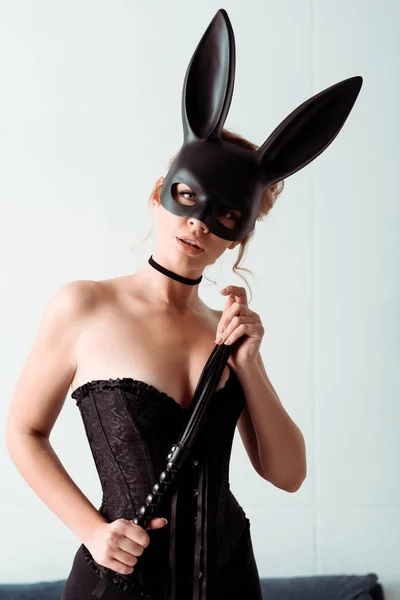 Dominante jeune femme en lapin masque et corset tenant fouet fouet et regardant caméra — Photo de stock