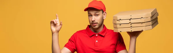Repartidor en uniforme rojo sosteniendo cajas de pizza y mostrando gesto de idea aislado en amarillo, tiro panorámico - foto de stock