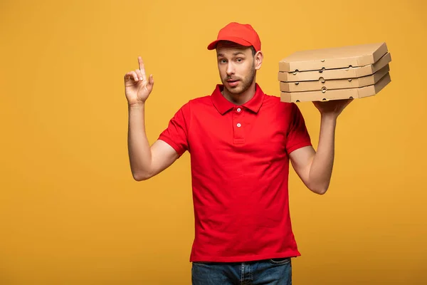 Repartidor en uniforme rojo sosteniendo cajas de pizza y mostrando gesto de idea aislado en amarillo - foto de stock
