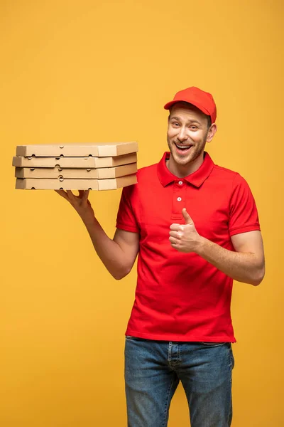 Feliz repartidor en uniforme rojo sosteniendo cajas de pizza y mostrando el pulgar hacia arriba aislado en amarillo - foto de stock