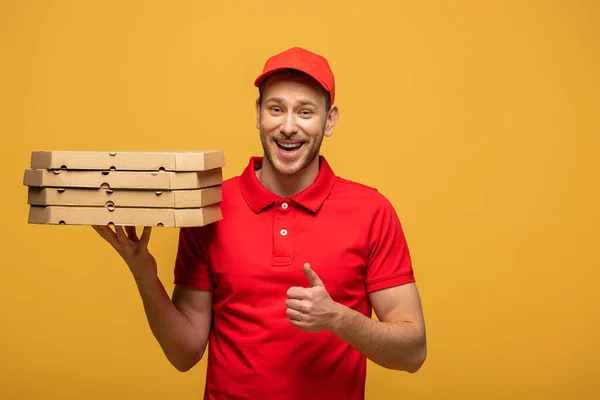 Felice uomo di consegna in uniforme rossa tenendo scatole di pizza e mostrando pollice in su isolato sul giallo — Foto stock