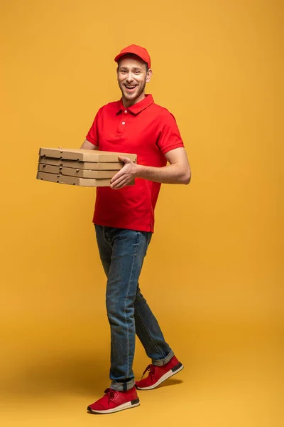 Vista completa de hombre entrega feliz en uniforme rojo caminando con cajas de pizza en amarillo - foto de stock