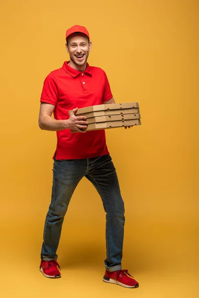 Visão de comprimento total de homem de entrega feliz em uniforme vermelho andando com caixas de pizza em amarelo — Fotografia de Stock