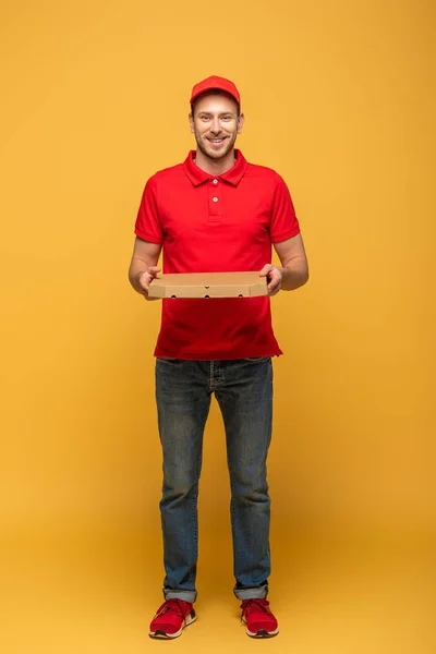 Vista completa de hombre de entrega feliz en uniforme rojo sosteniendo la caja de pizza en amarillo - foto de stock